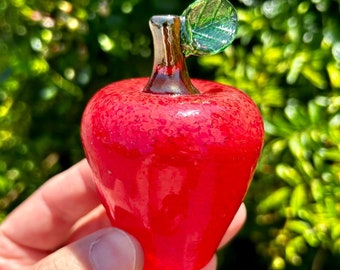 May include: A vibrant red glass apple with a textured surface. The apple features a brown stem and a green leaf. The decorative piece is held against a blurred green background.