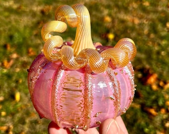 Può includere: Una zucca di vetro rosa con un gambo a spirale dorato. La zucca ha una superficie testurizzata ed è decorata con glitter dorato.