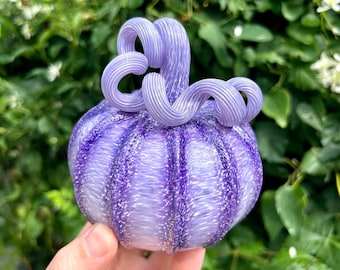 May include: A hand-blown glass pumpkin in shades of purple. The pumpkin has vertical ridges and a swirling stem. The glass is translucent, allowing light to pass through. The background is a blurred green with white flowers.