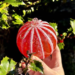 May include: A red glass Christmas ornament with white stripes and a frosted finish. The ornament is hanging from a gold wire.