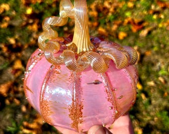 Può includere: Una zucca di vetro rosa con uno stelo a spirale dorato. La zucca ha una superficie testurizzata ed è decorata con brillantini dorati.