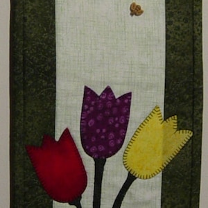 Puede incluir: Una pared de tela verde con un fondo blanco. El fondo presenta una mariposa azul, tres flores y una rana verde. Las flores son moradas, rojas y amarillas.