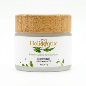 May include: A cylindrical glass jar with a light brown bamboo lid containing Holidentix remineralizing toothpowder. The white label features gold text, a tooth icon, green leaf illustrations, and states 2 oz | 56.7 g net weight.