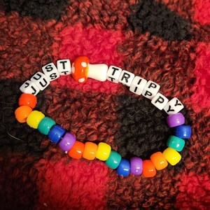 May include: A rainbow coloured beaded bracelet with the words "Just Trippy" spelled out in white beads. A red and white mushroom charm is in the centre of the bracelet.