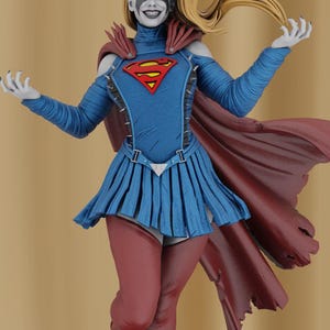 May include: A female action figure with a red and blue costume, a red cape, and a black and white face paint design. The figure is posed with arms outstretched and a mischievous smile.