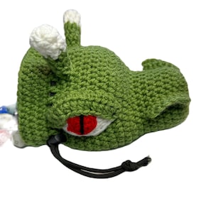 Puede incluir: Bolsa de monstruo de color verde hecha a crochet con un ojo rojo y detalles blancos. La bolsa tiene un cierre de cord&oacute;n y est&aacute; llena de dados de colores.