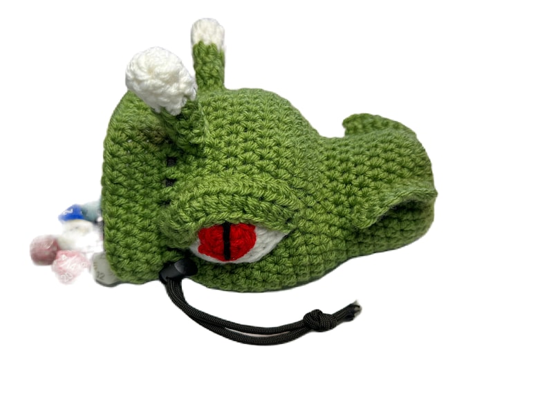 Puede incluir: Bolsa de monstruo de color verde hecha a crochet con un ojo rojo y detalles blancos. La bolsa tiene un cierre de cord&oacute;n y est&aacute; llena de dados de colores.