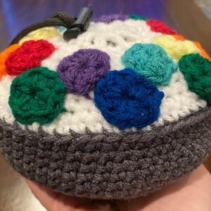 May include: A crocheted pouch with a white base and a gray bottom. The pouch is decorated with colorful crocheted circles in shades of red, orange, yellow, green, blue, purple, and white.