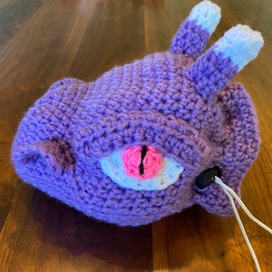 Puede incluir: Juguete de peluche de color morado tejido a crochet con orejas blancas y un ojo rosa. El juguete tiene un cord&oacute;n blanco.