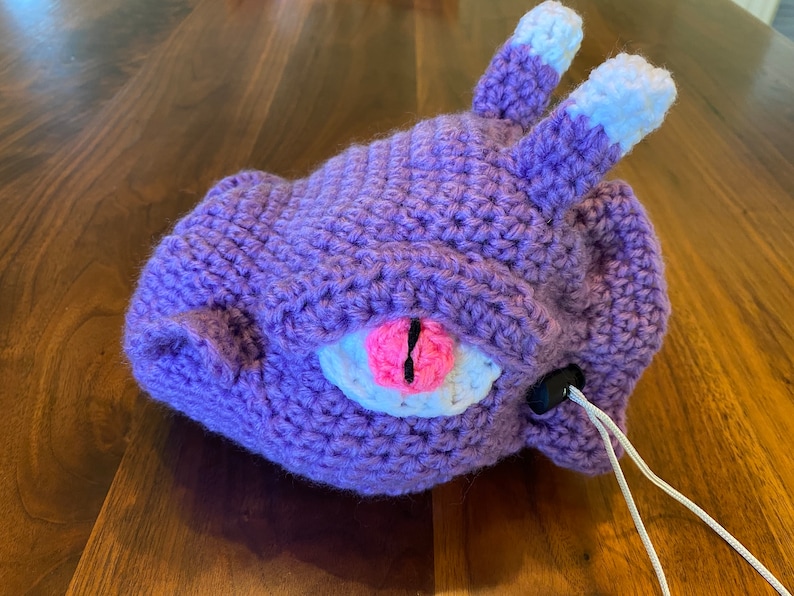 Puede incluir: Juguete de peluche de color morado tejido a crochet con orejas blancas y un ojo rosa. El juguete tiene un cord&oacute;n blanco.