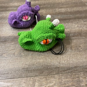 Puede incluir: Dos juguetes de peluche de crochet con forma de dragones con ojos grandes. Un drag&oacute;n es morado y el otro es verde.
