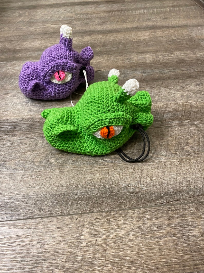 Puede incluir: Dos juguetes de peluche de crochet con forma de dragones con ojos grandes. Un drag&oacute;n es morado y el otro es verde.