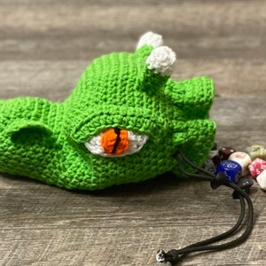 Puede incluir: Bolsa de dados de drag&oacute;n de crochet verde con un ojo blanco y una pupila naranja. La bolsa est&aacute; abierta y se derraman dados de colores.