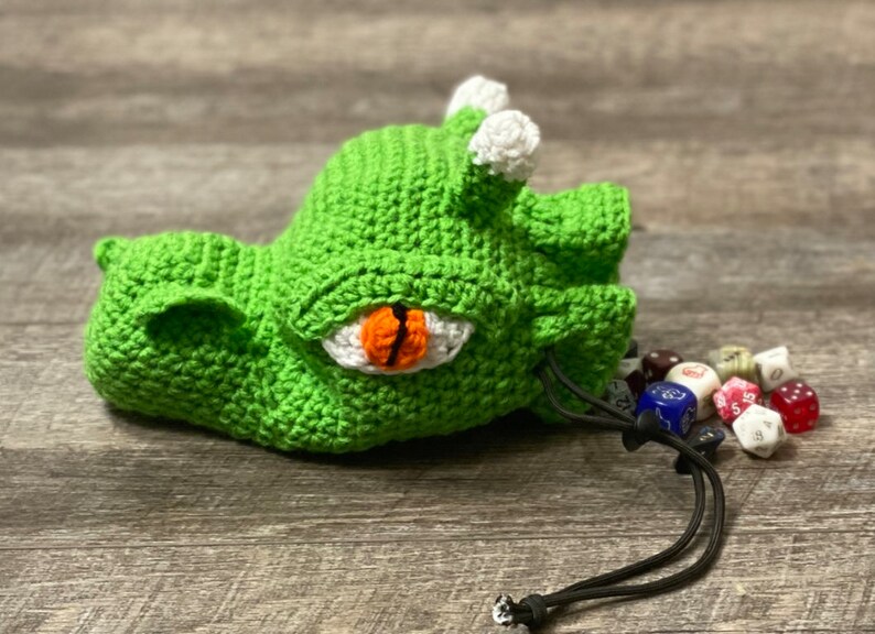 Puede incluir: Bolsa de dados de drag&oacute;n de crochet verde con un ojo blanco y una pupila naranja. La bolsa est&aacute; abierta y se derraman dados de colores.
