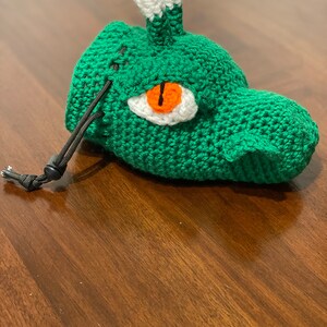 Puede incluir: Bolsa de dados de crochet verde con una cabeza de drag&oacute;n de crochet blanco y verde y un ojo naranja. La bolsa tiene un cord&oacute;n de ajuste negro.