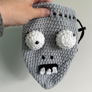 May include: A gray crocheted drawstring bag with a monster face. The monster has white eyes with black pupils, a black mouth, and white teeth.