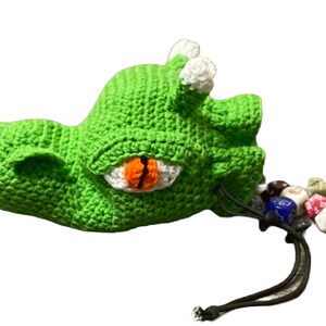 Puede incluir: Bolsa de dados de drag&oacute;n de crochet verde con un ojo blanco y una pupila naranja. La bolsa est&aacute; abierta y se derraman dados de colores.