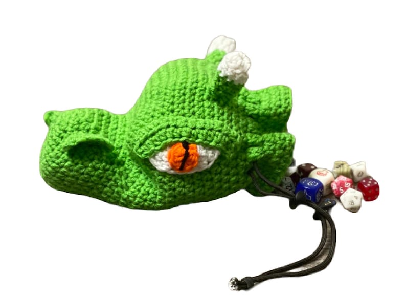 Puede incluir: Bolsa de dados de drag&oacute;n de crochet verde con un ojo blanco y una pupila naranja. La bolsa est&aacute; abierta y se derraman dados de colores.
