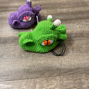 Puede incluir: Dos juguetes de peluche de crochet con forma de dragones con ojos grandes. Un drag&oacute;n es morado y el otro es verde.