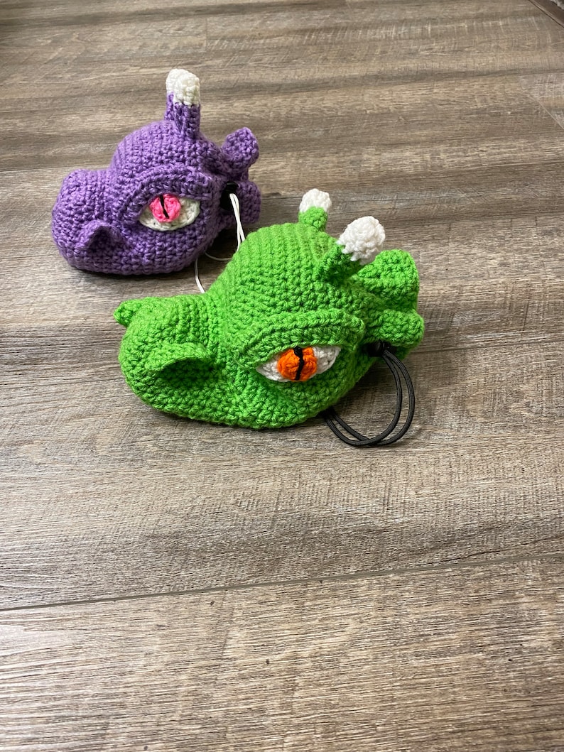 Puede incluir: Dos juguetes de peluche de crochet con forma de dragones con ojos grandes. Un drag&oacute;n es morado y el otro es verde.