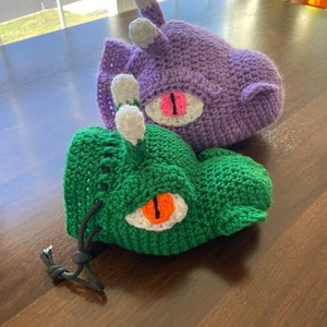 Puede incluir: Dos bolsas de dados de crochet, una verde y otra morada, con ojos blancos y naranjas. La bolsa verde tiene un cord&oacute;n de ajuste negro.