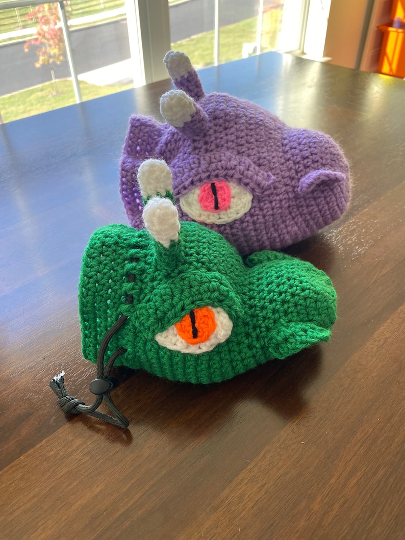 Puede incluir: Dos bolsas de dados de crochet, una verde y otra morada, con ojos blancos y naranjas. La bolsa verde tiene un cord&oacute;n de ajuste negro.