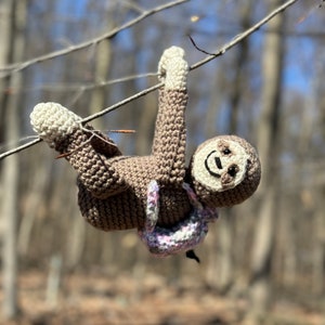 May include: A brown crocheted sloth hanging from a branch with a pink and white crocheted backpack. The sloth has a smiling face with black eyes.