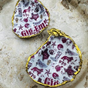 May include: Two oyster shell trinket dishes with gold gilded edges. The interior features a white background with maroon and gray graphics, including the text "Gig 'Em!" and "979", along with stars, boots, and footballs.