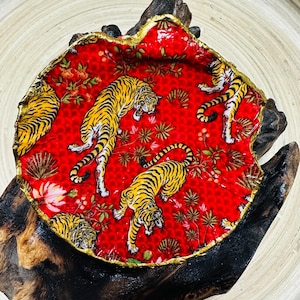 May include: A decorative red dish with a gold rim, featuring a repeating pattern of yellow and black tigers. The dish is set on a piece of dark wood, creating a striking contrast. The dish has a unique shape.