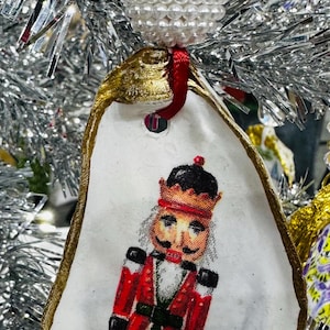 Op de afbeelding: Een kerstversiering gemaakt van een witte oesterschelp, met een geschilderde notenkrakersoldaat in een rood uniform. De schelp is afgewerkt met goudverf en bekroond met een parelparelcluster en rood lint om op te hangen.