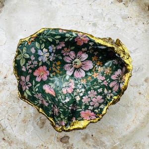May include: A decorative, floral-patterned trinket dish. The interior features a dark green background with pink, orange, and blue flowers. The rim is edged with gold. The dish is likely made from a natural shell.