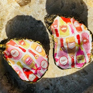 Puede incluir: Dos conchas de ostra con bordes pintados de oro. Las conchas están decoradas con un patrón de botellas de champán en rosa, rojo y amarillo. Las conchas están sobre una superficie marrón claro.