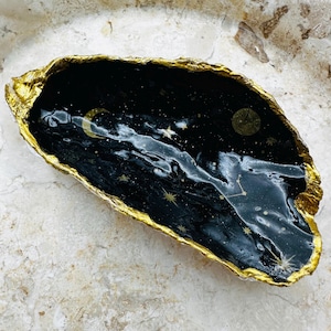 Pode incluir: Uma tigela decorativa preta e dourada com um design celestial. O interior apresenta uma cena de céu noturno com uma lua crescente, estrelas e constelações. A tigela tem uma borda dourada texturizada.
