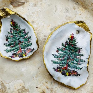 May include: Two oyster shell ornaments with a Christmas tree design. The shells have a gold-painted edge and a white interior. The Christmas tree is green with red, yellow, and blue ornaments and presents at the base.