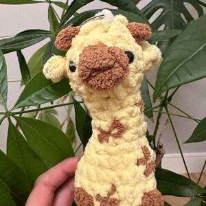 Puede incluir: Un juguete de peluche de jirafa amarillo y marrón con nariz marrón y ojos negros. La jirafa sostiene un anillo plateado en su boca.