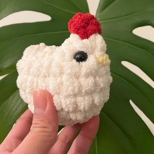 Puede incluir: Un juguete de peluche de pollo blanco tejido a crochet con un pomp&oacute;n rojo en la cabeza. El pollo tiene ojos negros y un pico amarillo.