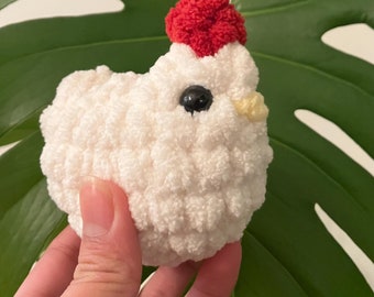 May include: A white crocheted chicken plush toy with a red pom-pom on its head. The chicken has black eyes and a yellow beak.