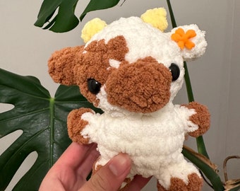 May include: A white and brown crocheted cow plush toy with a yellow flower on its ear. The toy has black eyes and a brown nose.