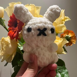 Puede incluir: Peluche de conejo blanco de crochet con ojos negros y una X negra en la nariz. El conejo está siendo sostenido en una mano contra un fondo de rosas amarillas y flores rojas.