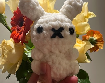 May include: White crocheted bunny rabbit plush toy with black eyes and a black X on its nose. The bunny is being held in a hand against a background of yellow roses and red flowers.