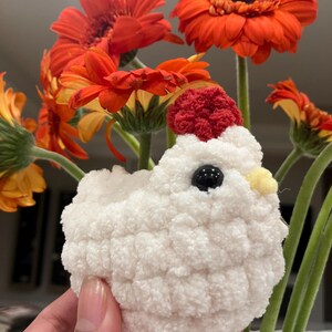 Puede incluir: Un juguete de pollo blanco de crochet con una cresta roja y un pico amarillo. El pollo est&aacute; sentado en una mano frente a un ramo de margaritas naranja.