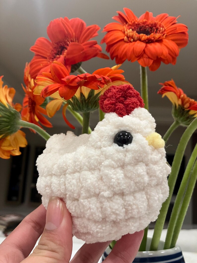 Puede incluir: Un juguete de pollo blanco de crochet con una cresta roja y un pico amarillo. El pollo est&aacute; sentado en una mano frente a un ramo de margaritas naranja.