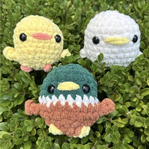Puede incluir: Tres animales de peluche de crochet con forma de patos. Los patos son amarillos, blancos y verde y marrón. Los patos están sentados en una cama de hojas verdes.