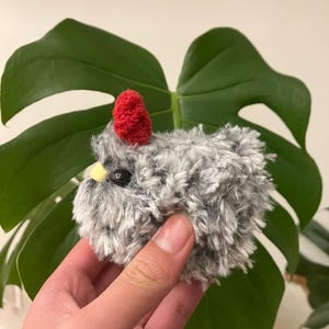 Puede incluir: Un peque&ntilde;o pollo de crochet gris y blanco con un pomp&oacute;n rojo en la cabeza. El pollo tiene ojos negros y un pico amarillo.