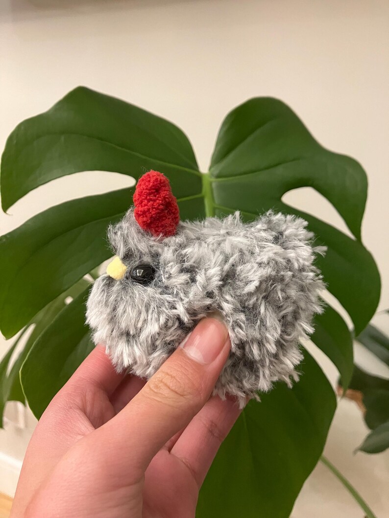 Puede incluir: Un peque&ntilde;o pollo de crochet gris y blanco con un pomp&oacute;n rojo en la cabeza. El pollo tiene ojos negros y un pico amarillo.
