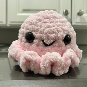 Puede incluir: Un juguete de peluche de pulpo rosa tejido a crochet con ojos negros y una boca sonriente.