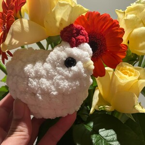 Puede incluir: Un juguete de pollo blanco de crochet con una cresta roja y ojos negros, rodeado de flores amarillas y rojas.