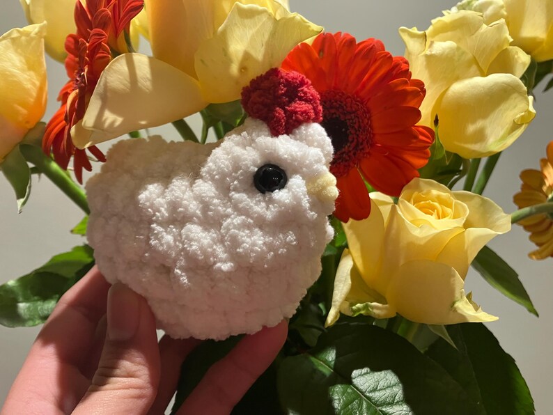 Puede incluir: Un juguete de pollo blanco de crochet con una cresta roja y ojos negros, rodeado de flores amarillas y rojas.