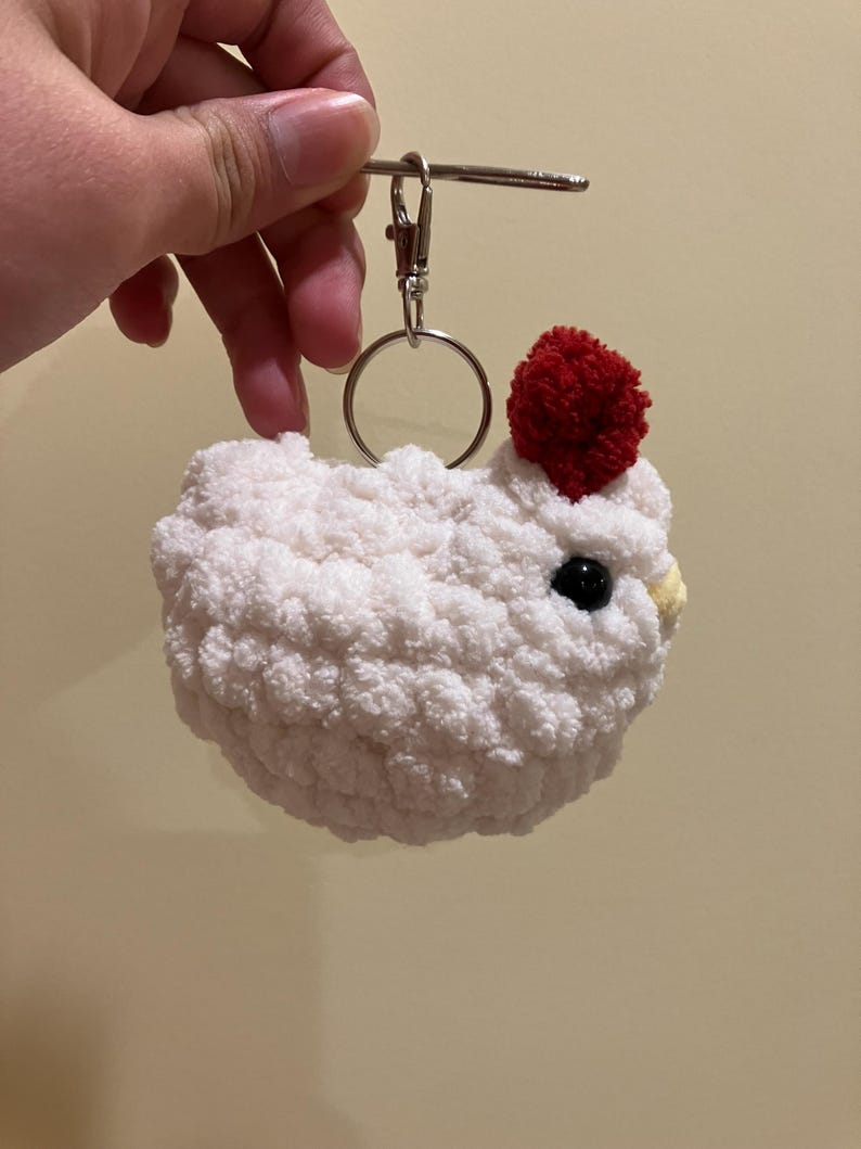 Puede incluir: Llavero de pollo blanco tejido a crochet con un pomp&oacute;n rojo en la cabeza. El llavero tiene un anillo de metal plateado y un cierre de metal plateado.
