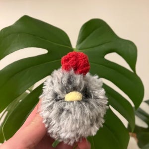 Puede incluir: Un peque&ntilde;o pollo de crochet gris y blanco con una cresta roja de crochet. El pollo tiene un pico amarillo y ojos negros.